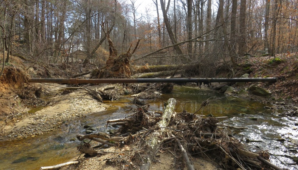 Aerial crossing of a sanitary sewer line caused by stream degradation and expansion. The crossing is at risk of causing debris jams.