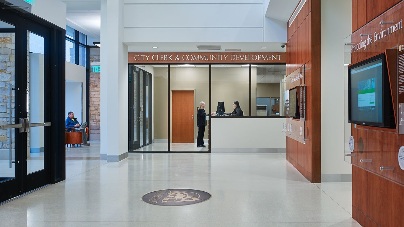 The single public entry in this combined facility features a dramatic, light-filled two-story lobby with prairie-style inspired pendant light fixtures, wood paneled signage and display walls, and seating areas with views out to the civic lawn.