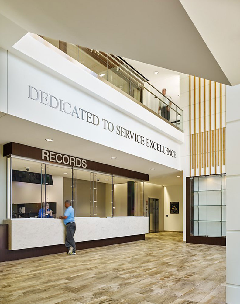 The two-story, light-filled public lobby features warm natural colors, wood accents, a display case, and ballistic-rated glazing and counter details at the records window.