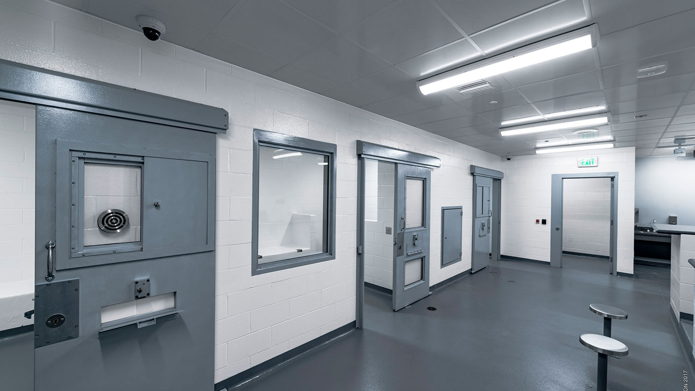 The temporary holding facility includes sliding cell doors, security ceilings, and an elevated booking counter platform with direct line of site into all cells.